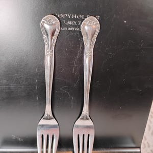 May include: Two silver forks with ornate handles are displayed against a dark background. The handles have a floral design at the top. The word "Liberty" is embossed at the top of the background.
