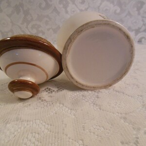 Peut inclure: Un pot en c&eacute;ramique blanc avec un couvercle et une base marron. Le couvercle a un bord marron circulaire et un centre beige. Le pot est ouvert, r&eacute;v&eacute;lant son int&eacute;rieur. Le pot et le couvercle sont sur une surface en dentelle blanche.