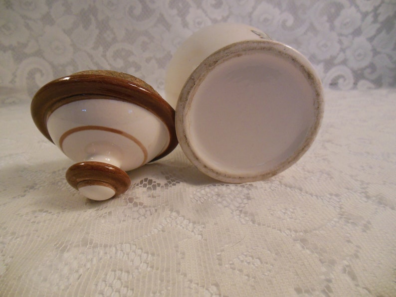 Peut inclure: Un pot en c&eacute;ramique blanc avec un couvercle et une base marron. Le couvercle a un bord marron circulaire et un centre beige. Le pot est ouvert, r&eacute;v&eacute;lant son int&eacute;rieur. Le pot et le couvercle sont sur une surface en dentelle blanche.