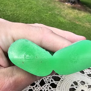 Puede incluir: Un par de zuecos de juguete de pl&aacute;stico verde brillante, sostenidos en una mano, con texto en relieve. Los zuecos descansan sobre un tapete crema de tejido abierto. El fondo muestra una zona de c&eacute;sped y &aacute;rboles.