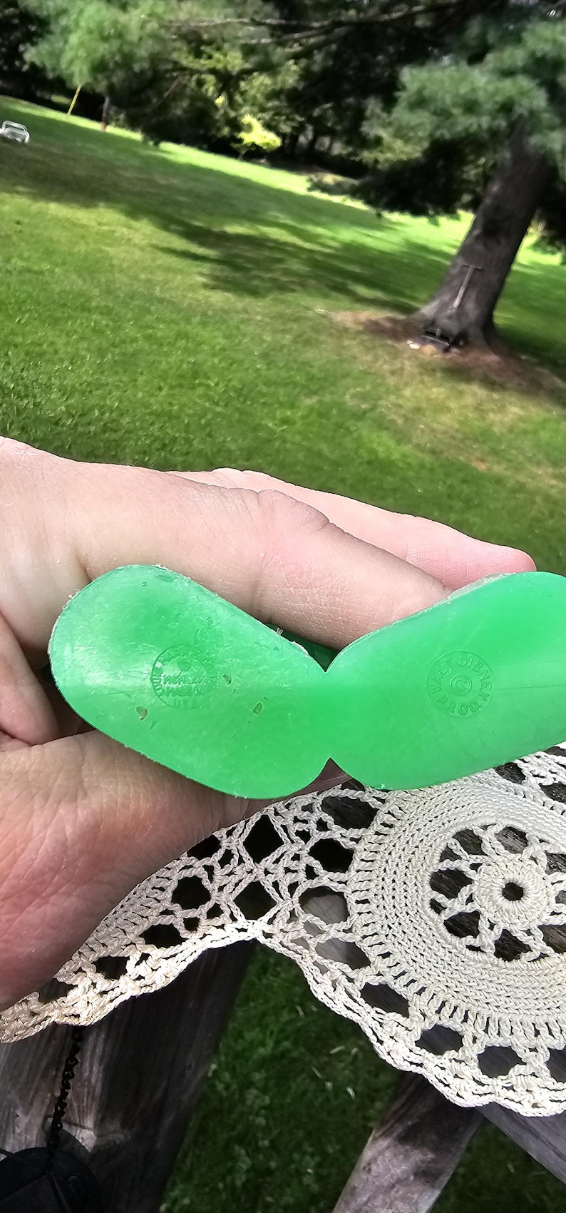 Puede incluir: Un par de zuecos de juguete de pl&aacute;stico verde brillante, sostenidos en una mano, con texto en relieve. Los zuecos descansan sobre un tapete crema de tejido abierto. El fondo muestra una zona de c&eacute;sped y &aacute;rboles.