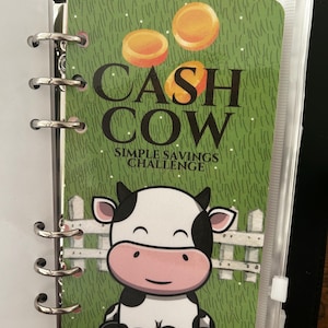 May include: A green and white cash cow savings challenge tracker with a cartoon cow sitting on a green field with a white picket fence. The tracker has the words "CASH COW" and "SIMPLE SAVINGS CHALLENGE" printed on it.