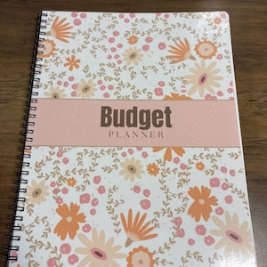 May include: A spiral-bound budget planner with a floral design. The cover features a white background with orange, peach, and light brown flowers and leaves. The title "Budget Planner" is displayed in dark brown letters on a pink band.