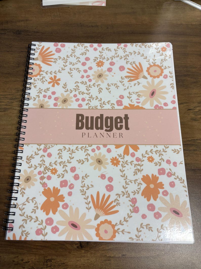 May include: A spiral-bound budget planner with a floral design. The cover features a white background with orange, peach, and light brown flowers and leaves. The title "Budget Planner" is displayed in dark brown letters on a pink band.