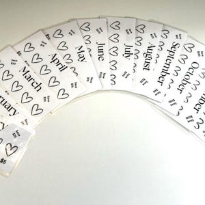 May include: Twelve white cards with black hearts and the months of the year written in black text. Each card has a different month written on it, starting with January and ending with December. The cards are arranged in a semi-circle, with the January card at the bottom and the December card at the top.