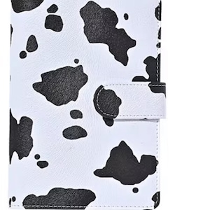 May include: A white planner featuring a black cow print design. The planner has a flap closure with a press stud. It is designed to hold inserts and has a tabbed section at the bottom. A stylish way to stay organised.