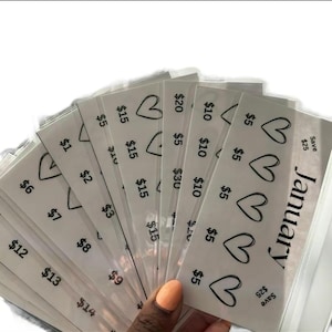 May include: A collection of white paper savings envelopes with black text and heart designs. Each envelope is labelled with a monetary value, ranging from &pound;1 to &pound;30. The word "January" is printed on one envelope.