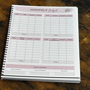 May include: A spiral-bound notebook with a "MONTHLY Budget" title in script. The pages are white with pink accents and include sections for debt, savings, and expenses. The notebook is open on a dark wood surface.