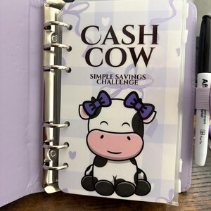 May include: A lilac binder with a "Cash Cow Simple Savings Challenge" cover. The cover features a cartoon cow with purple bows. A black marker with a purple band is visible on the right side.