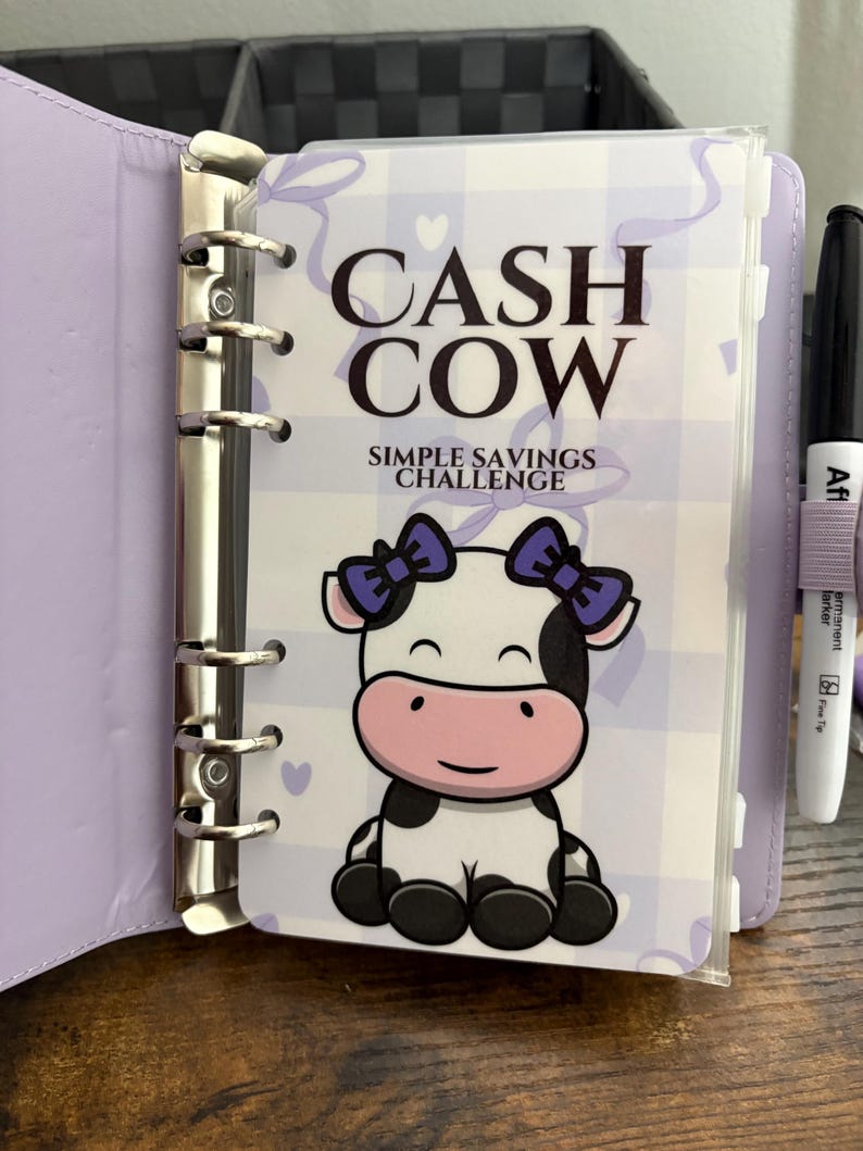 May include: A lilac binder with a "Cash Cow Simple Savings Challenge" cover. The cover features a cartoon cow with purple bows. A black marker with a purple band is visible on the right side.