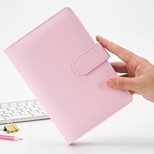 May include: A pink leather bound planner with a flap closure and a strap to keep it closed. The planner is held in a hand.