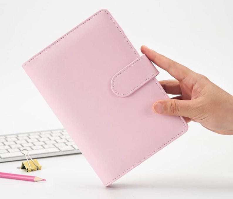 May include: A pink leather bound planner with a flap closure and a strap to keep it closed. The planner is held in a hand.