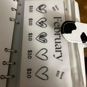 May include: A white binder with silver rings, containing a clear plastic pocket with a February savings tracker. The tracker displays heart graphics and text indicating &pound;10 savings per heart, with a &pound;50 savings goal.