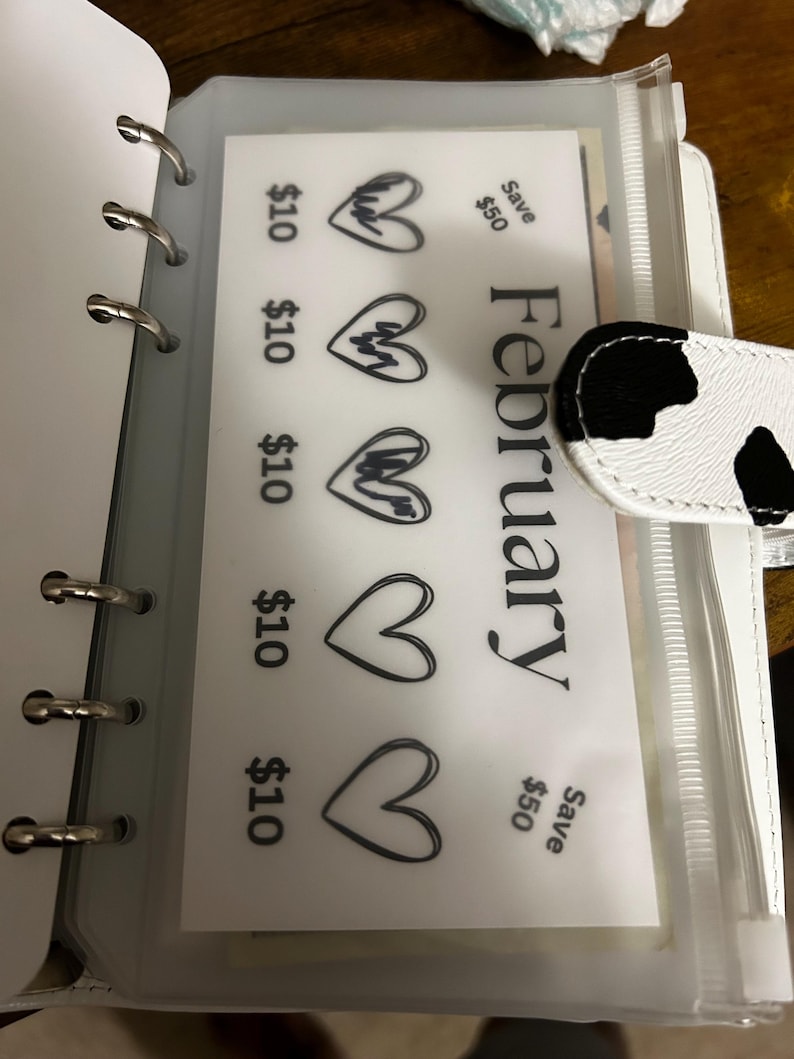 May include: A white binder with silver rings, containing a clear plastic pocket with a February savings tracker. The tracker displays heart graphics and text indicating &pound;10 savings per heart, with a &pound;50 savings goal.