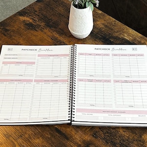 May include: Open spiral-bound planner with the title "PAYCHECK Breakdown" in black script. The pages are white with pink accents and include sections for income, expenses, and budget summaries. A white vase with purple flowers sits in the background.