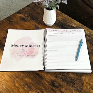 May include: An open spiral-bound notebook with the title "Money Mindset Reflection on Financial Challenges" and a "Financial Therapy Worksheet" is on a wooden table. A blue pen rests on the open page. A small white vase with purple flowers sits nearby.