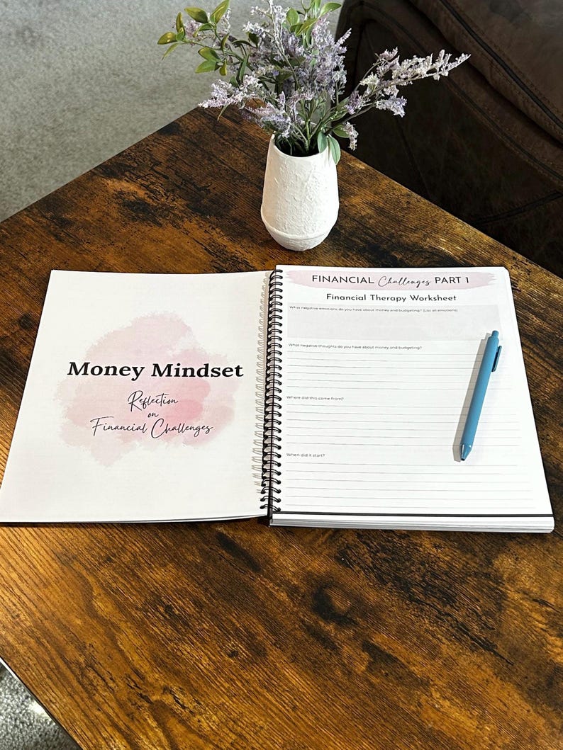 May include: An open spiral-bound notebook with the title "Money Mindset Reflection on Financial Challenges" and a "Financial Therapy Worksheet" is on a wooden table. A blue pen rests on the open page. A small white vase with purple flowers sits nearby.