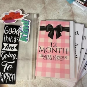 May include: A pink and white checkered card with a black bow and the text "12 Month Simple Savings Challenge". The card is part of a stack of cards with the text "Save $75" and "Save $105" visible.