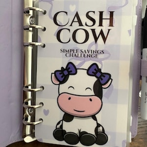 May include: A lilac binder with silver rings, open to a page reading "CASH COW SIMPLE SAVINGS CHALLENGE." The page shows a cartoon cow with purple bows and a pink nose, on a checkered background.