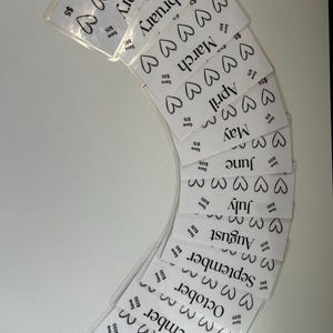 May include: Twelve white cards with black hearts and the months of the year printed on them. Each card has a different amount of hearts and a different dollar amount printed on it. The cards are arranged in a semi-circle.