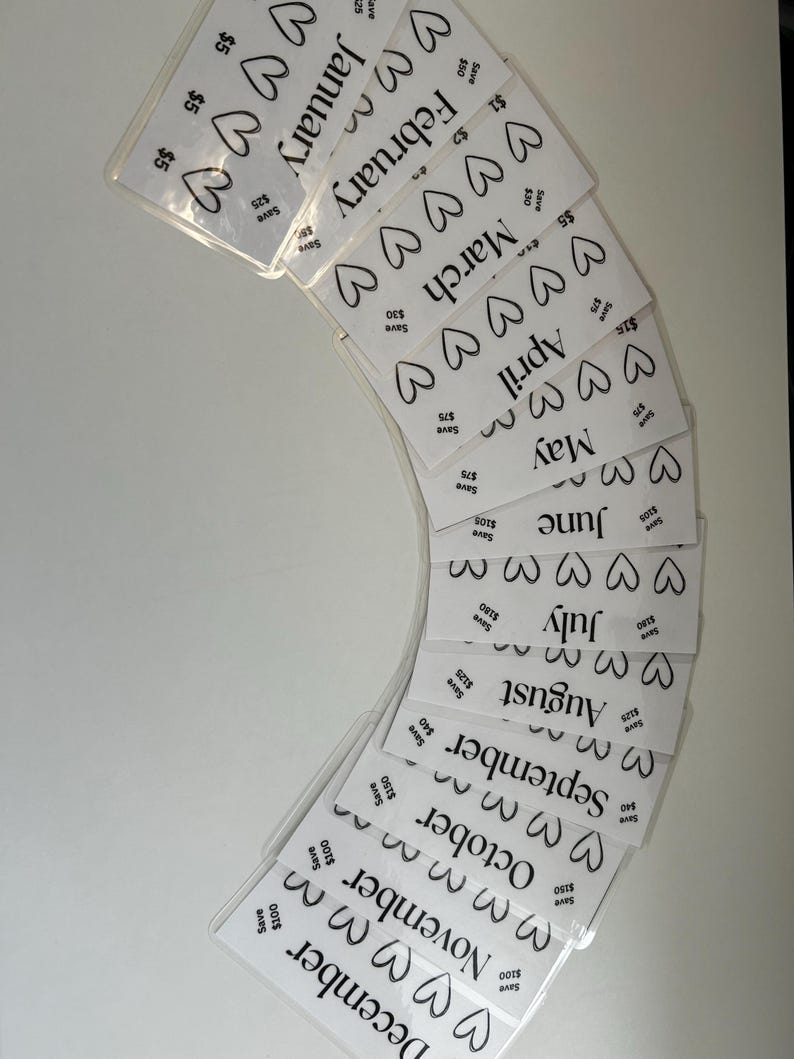 May include: Twelve white cards with black hearts and the months of the year printed on them. Each card has a different amount of hearts and a different dollar amount printed on it. The cards are arranged in a semi-circle.