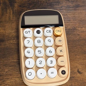 Puede incluir: Una calculadora beige con una pantalla negra y botones blancos y beige. La calculadora tiene forma rectangular con bordes redondeados. Los botones incluyen números, símbolos matemáticos y teclas de función.