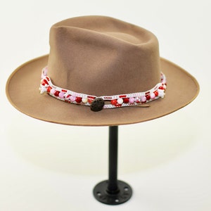 May include: A brown felt fedora hat with a white and red beaded band. The hat is on a black stand.