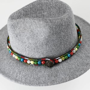 May include: A gray felt fedora hat with a colorful beaded band. The band features a variety of colors, including red, green, blue, and yellow. The hat has a wide brim and a button on the top.