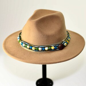 May include: A tan fedora hat with a decorative band of colorful beads. The band features blue, yellow, green, and white beads, and a brown button. The hat is displayed on a black stand against a white background.