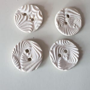 May include: Four white ceramic buttons with a textured, swirling design and two holes each. The buttons are round and have a glossy finish. Decorative white accents are in the background.