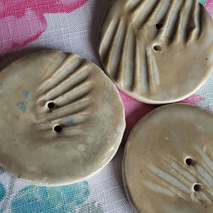 May include: Three ceramic buttons with a light brown glaze and white lines. The buttons have a raised design and a hole in the center.