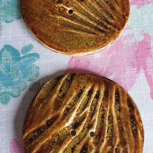 May include: Two brown ceramic buttons with a textured, wavy design and two holes each. The buttons are on a white and pink floral fabric.