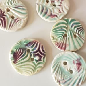 May include: A collection of five ceramic buttons with a cream-colored base. Each button features a unique embossed design with green and burgundy accents. The buttons have two holes for sewing.