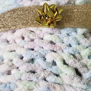 May include: A handmade, chunky knit blanket in pastel colors of blue, pink, and green. A gold glitter ribbon is wrapped around the blanket, and a gold bow is placed on top. The blanket is soft and cozy.
