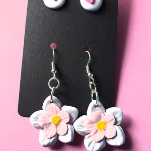 May include: A pair of white flower earrings with pink centers and yellow accents. The earrings are on a black card with two white stud earrings with pink hearts.