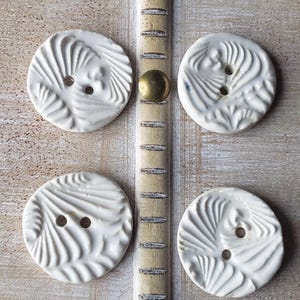 May include: Four white ceramic buttons with a textured, swirling design. The buttons have two holes for sewing and are arranged on a light-colored wooden surface with brass accents. Decorative sewing notions.