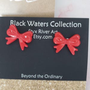 May include: A pair of bright red bow-shaped stud earrings. The earrings are displayed on a black card with the text "Black Waters Collection" and "Beyond the Ordinary". The background is a light brown and pink surface.
