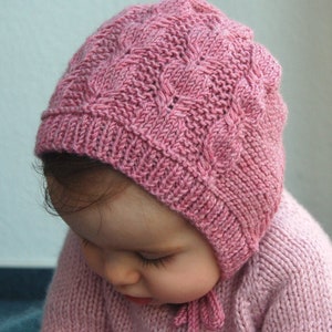 Könnte beinhalten: Eine rosa gestrickte Babyhaube mit einem Zopfmuster. Die Haube hat eine spitze Krone und bindet unter dem Kinn.