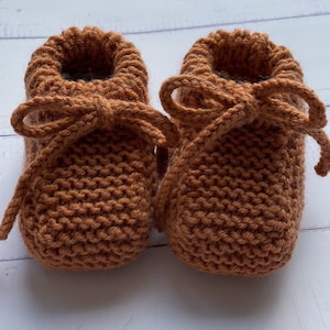 Pode incluir: Um par de sapatinhos de bebé de malha castanho com um laço amarrado em cada um. Os sapatinhos são feitos de um fio macio e texturizado.