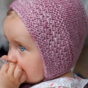 Könnte beinhalten: Nahaufnahme eines Babys, das eine rosa gestrickte Mütze mit einem strukturierten Muster trägt. Die Mütze bedeckt die Ohren und die Stirn des Babys.