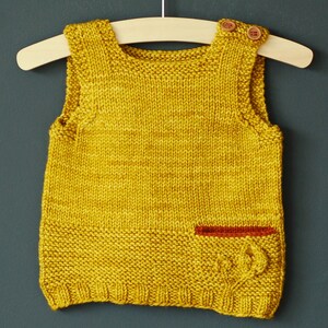 May include: A yellow knitted sleeveless vest with a red pocket detail. The vest is hanging on a wooden hanger against a dark green background.