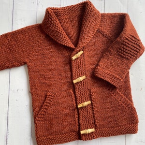 May include: A rust-colored knitted cardigan with a shawl collar and wooden toggle closures. The cardigan has two patch pockets and a ribbed knit pattern.