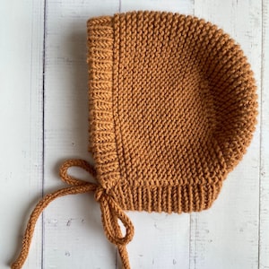 May include: A brown knitted baby bonnet with a tie. The bonnet is made of a soft, textured yarn.