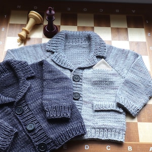 May include: Two knitted cardigans with button closures, one in a light gray color with a pocket and the other in a dark gray color. The cardigans are laid out on a wooden surface with a chessboard pattern.