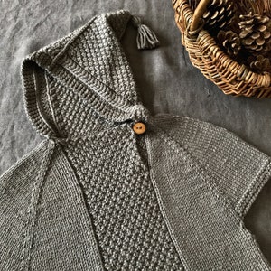Op de afbeelding: Een grijze gebreide cape met capuchon, een houten knoop en een kwast op de capuchon.