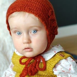 Könnte beinhalten: Ein Baby trägt eine gestrickte orange Mütze mit einem gestrickten gelben Pullover und einem weißen Hemd mit Blumenmuster.