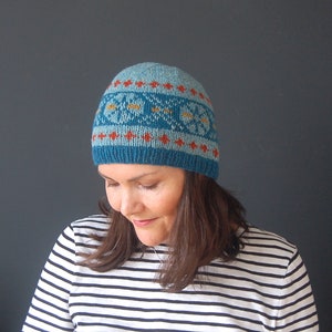 May include: A blue knitted hat with a geometric pattern in orange, yellow, and white. The hat is being worn by a person with brown hair.