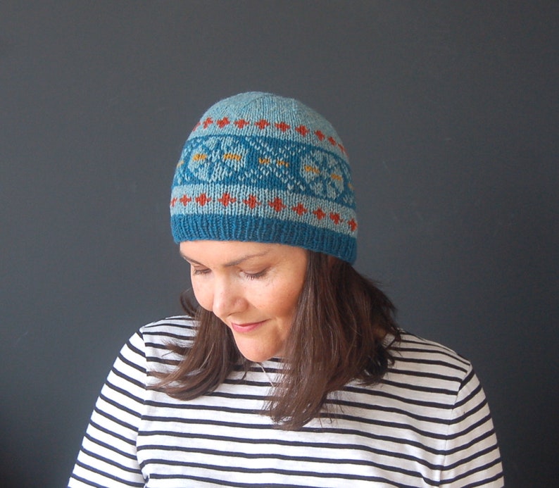 May include: A blue knitted hat with a geometric pattern in orange, yellow, and white. The hat is being worn by a person with brown hair.
