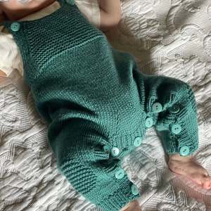 Puede incluir: Un mono de punto de color verde azulado para bebé con botones de color aguamarina. El mono está hecho de un tejido de punto suave y texturizado.