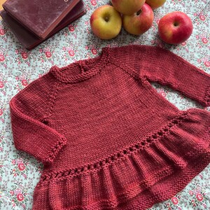 May include: A maroon knitted sweater with a ruffled hem. The sweater is made of a soft, textured yarn and has a relaxed fit.